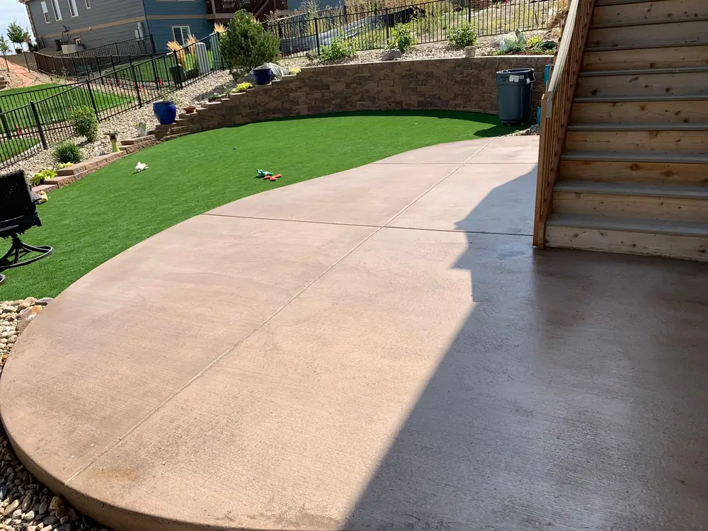 Curved stamped concrete patio beside artificial turf and deck stairs