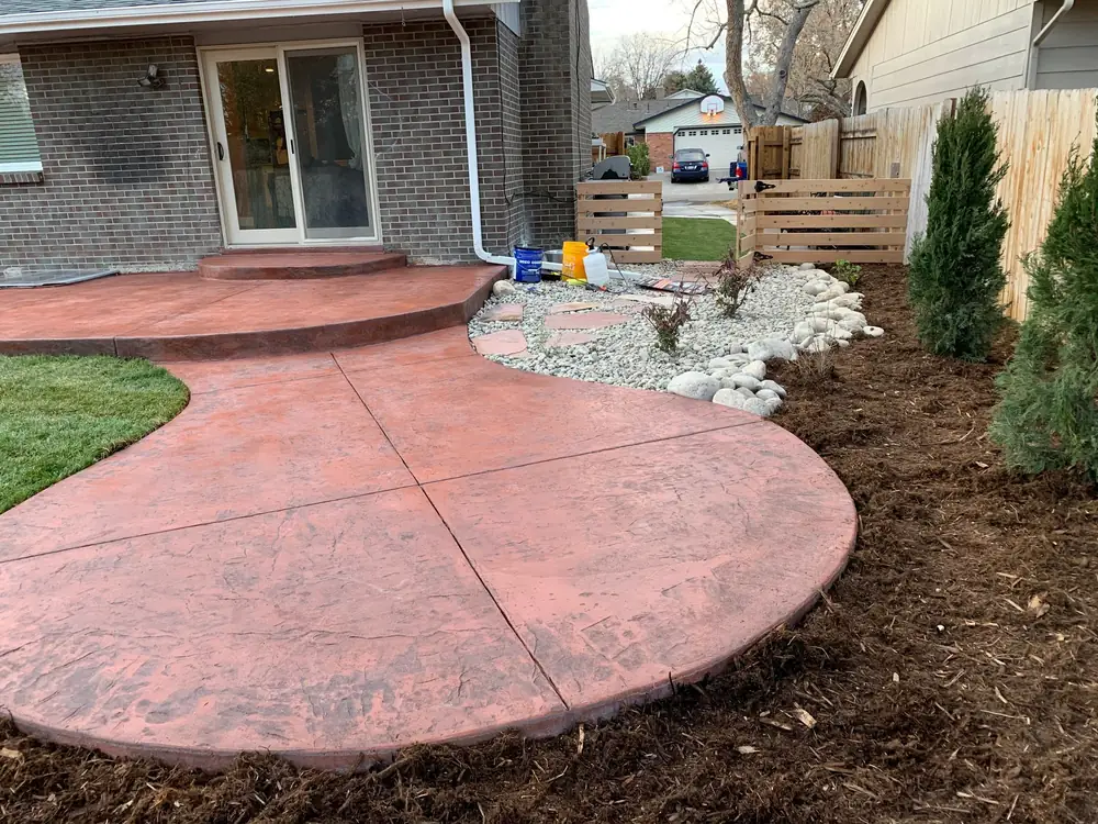 Red stamped concrete patio edged with river rock and young evergreens